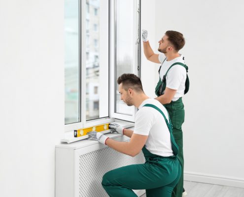 Two men in green overalls work on installing a window in a white room.