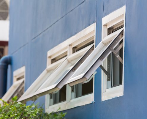 A close up of a blue wall on the exterior of a home, with several awning windows open in a line. The top of a shrub can be seen in the bottom left corner.