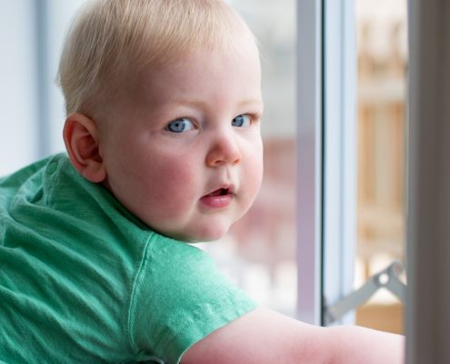 A baby boy looks at the camera as he is perched behind a child proof window.