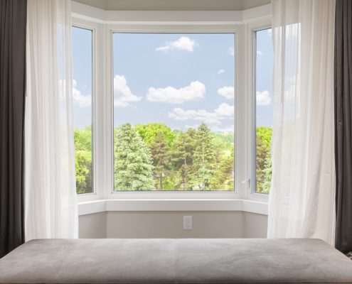 A closeup image of a bay window in a bedroom. You can see the gray wallpaper, white curtains, and the trees and skyline outside the house.