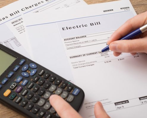 A closeup of a calculator next to a stack of papers on a desk. A person's hand holding a pen hovers above a sheet of paper that reads "electric bill".