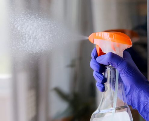 A purple-gloved hand squeezes down on a clear squirt bottle with an orange top to clean the clear glass in front of it. You can see the cleaning liquid shooting from the end of the spray bottle.