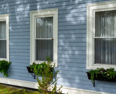 A series of three double hung windows as viewed from the outside of a blue house. You can see a windowsill with plants underneath each window.