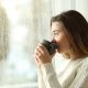 A woman sips out of a brown coffee mug while looking out of a window. You can see a plethora of raindrops running down the pane.